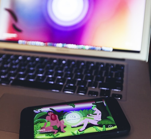 A smartphone displaying an app or game with cartoonish characters and greenery is resting on a laptop keyboard. The laptop screen shows a blurred, colorful background with a circular design.